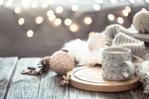 Coffee cup over Christmas lights bokeh in home on wooden table with sweater on a background and decorations. Winter mood, holiday decoration, magic Christmas.